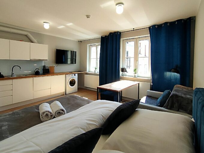 A modern studio living area with a white kitchen along the left, a washing machine, a wall-mounted TV, and a wooden table by large windows with blue curtains.