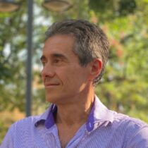 Portrait of a middle-aged man in a purple striped shirt, looking to the left, outdoors with greenery in the background.